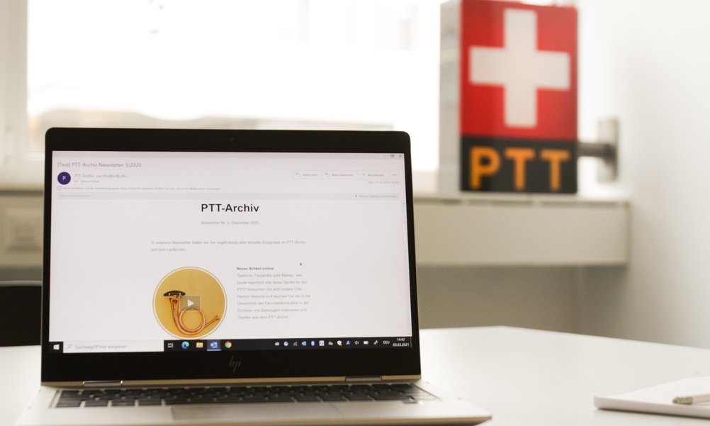 Auf einem Laptop ist ein Newsletter des PTT-Archives zu sehen. Im Hintergrund steht ein PTT-Logo als Leuchtkasten.