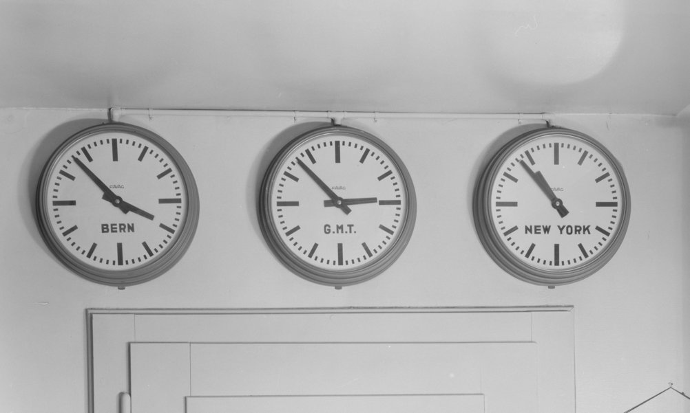 Trois horloges sur un mur indiquent des fuseaux horaires différents : Berne, Greenwich Mean Time et New York.