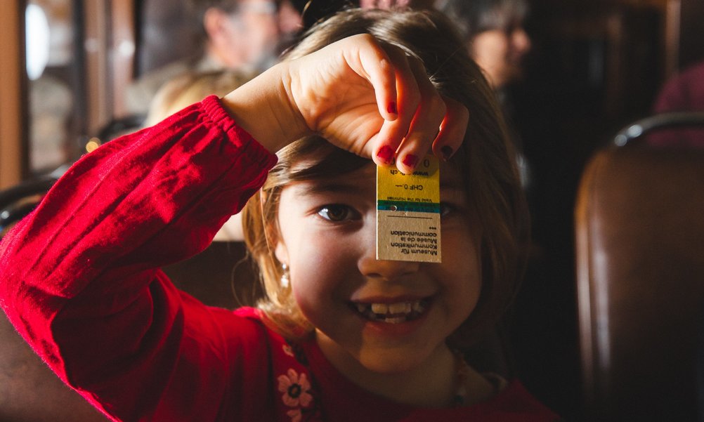 Une petite fille en robe rouge est assise dans un car postal historique et tient un ticket en carton devant son visage. 