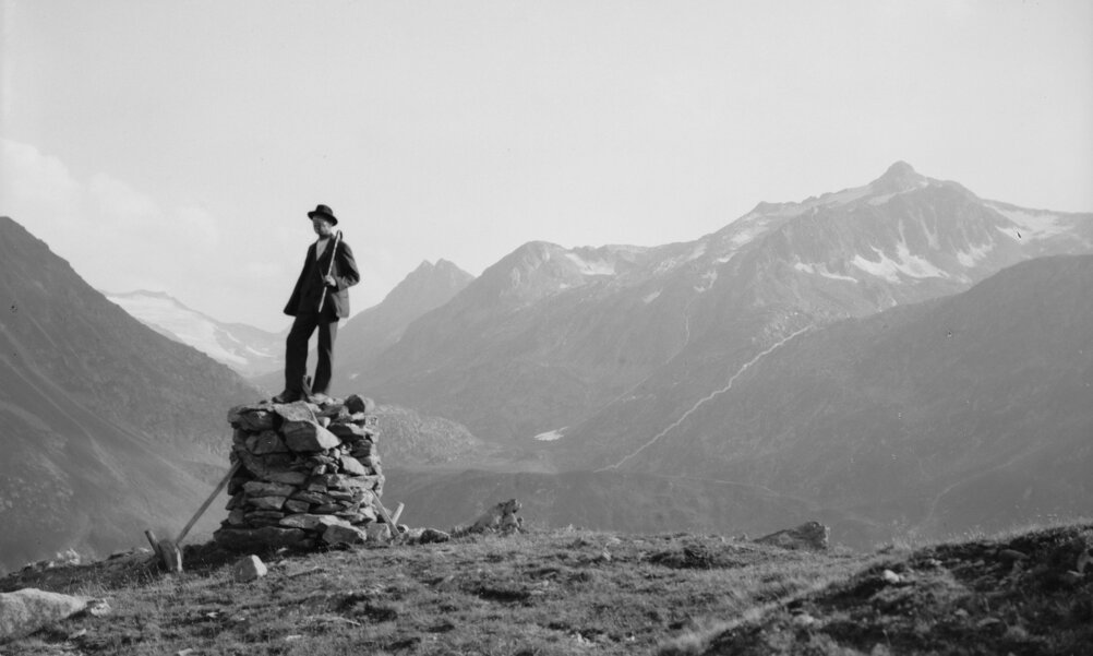 Ein schwarz-weiss-Bild aus den Bergen. Auf einem aufgemauerten Sockel aus Steinen steht ein Mann mit Stock und blickt in die Ferne. Im Hintergrund Berge.