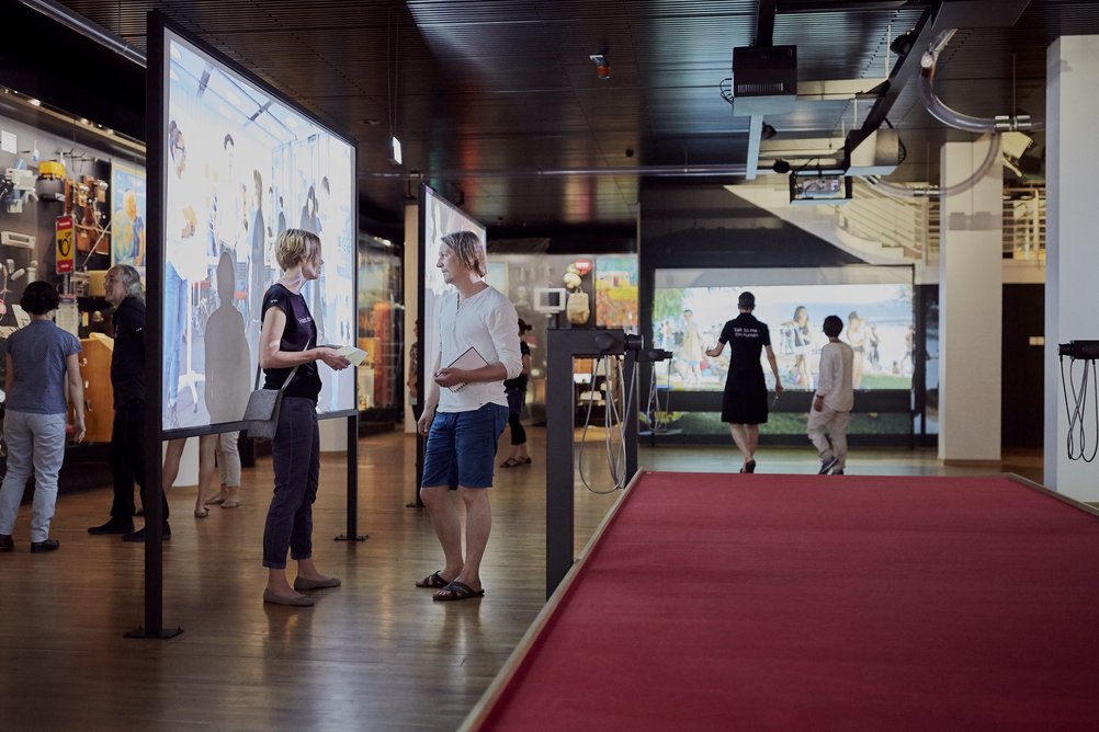 Blick in die Kernausstellung des Museums für Kommunikation: Um einen roten Teppich gruppieren sich Videoscreens, Objekte und Menschen, die sich unterhalten.