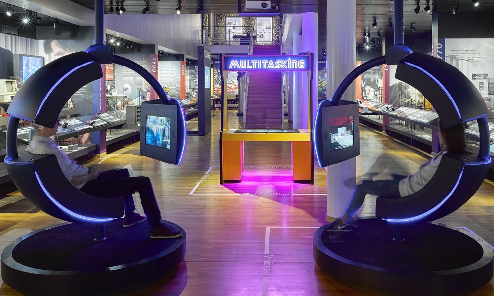 Ein Blick in die Zone Change in der Kernausstellung des Museums für Kommunikation. Im Vordergrund sind zwei futuristische Drehstühle zu sehen, im Hintergrund eine Spielstation.