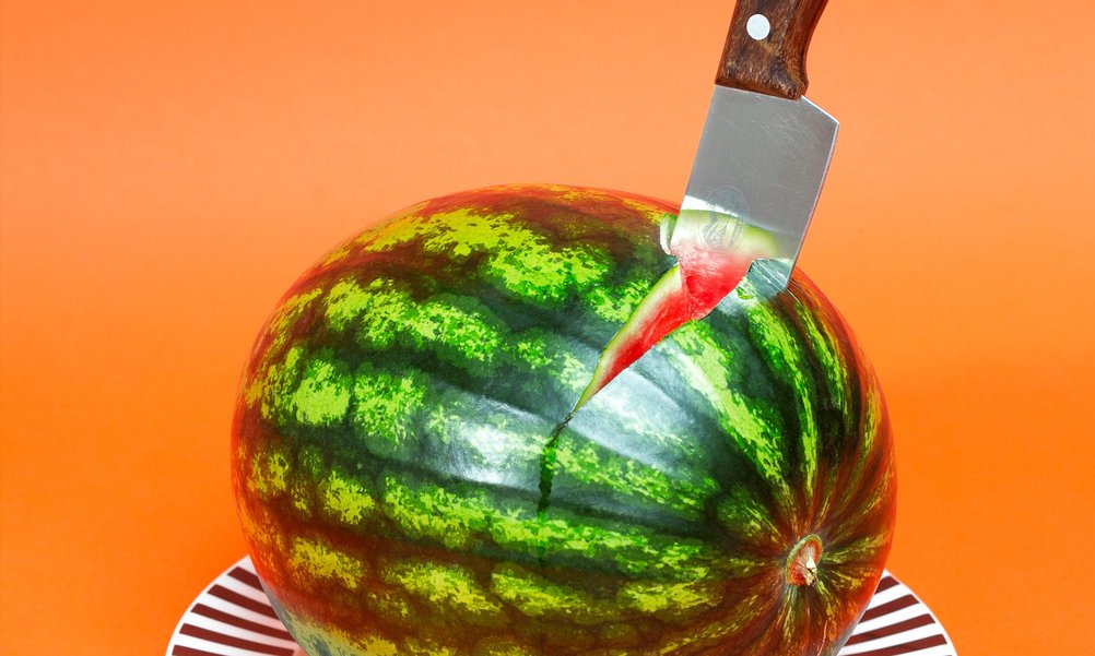 Eine grüne Wassermelone liegt auf einem Teller, darin steckt ein Messer.