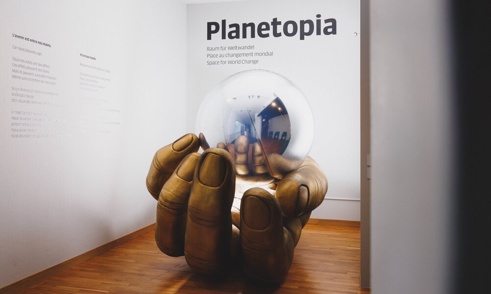 Eingang zur Ausstellung Planetopia. Eine überdimensionale goldene Hand hält eine silberne Spiegelkugel, darüber der Schriftzug Planetopia.