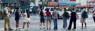 Eine vielfältige Gruppe von Menschen steht an einem New Yorker Strassenrand mit dem Rücken zur Kamera. Auf der anderen Strassenseite Häuser mit Reklamen und ein Haus im Baugerüst. - vergrösserte Ansicht