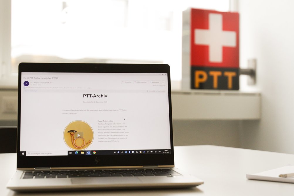 Un bulletin d'information des archives des PTT est visible sur un ordinateur portable. À l'arrière-plan, on voit le logo des PTT sous la forme d'une boîte lumineuse.