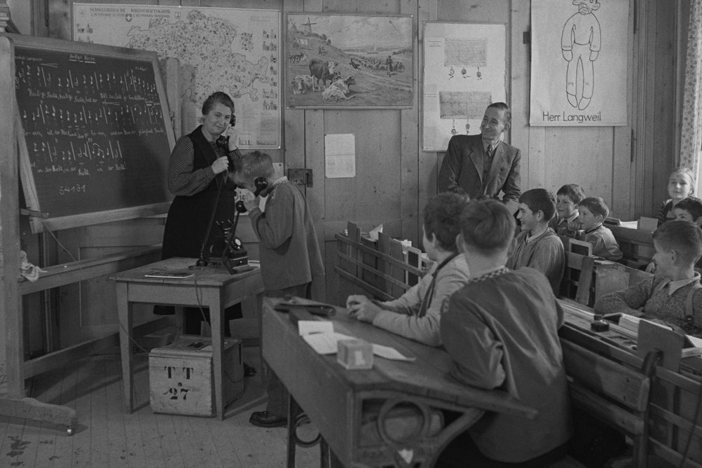 Ein historisches Bild zeigt wie eine Lehrerin mit einem Schüler vor der ganzen Klasse telefoniert.