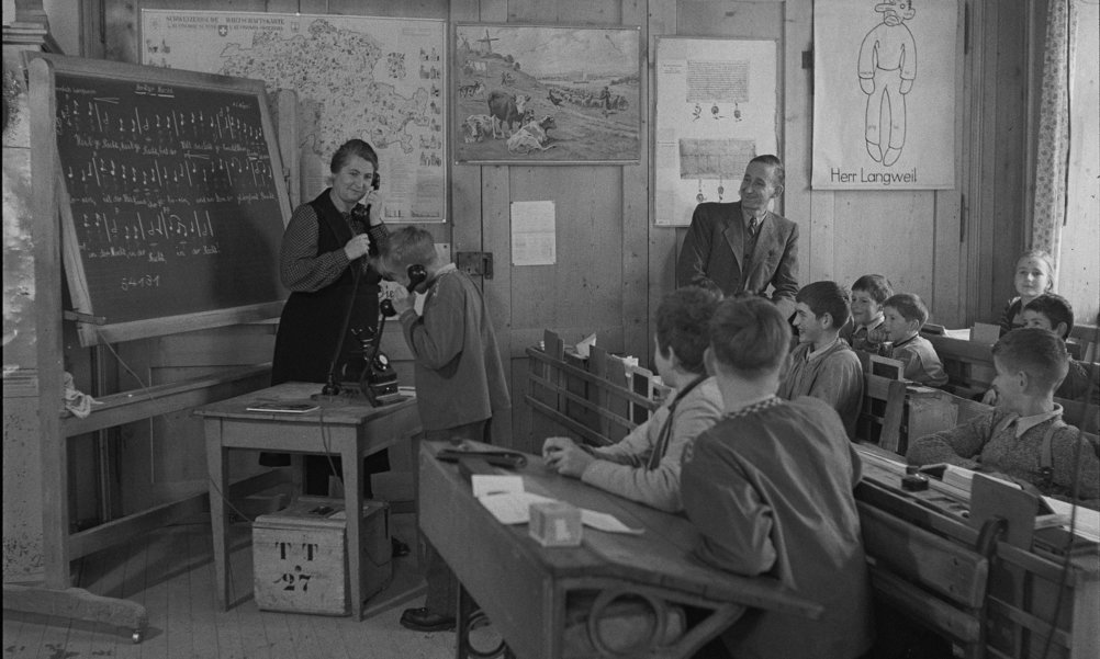 Ein historisches Bild zeigt wie eine Lehrerin mit einem Schüler vor der ganzen Klasse telefoniert.