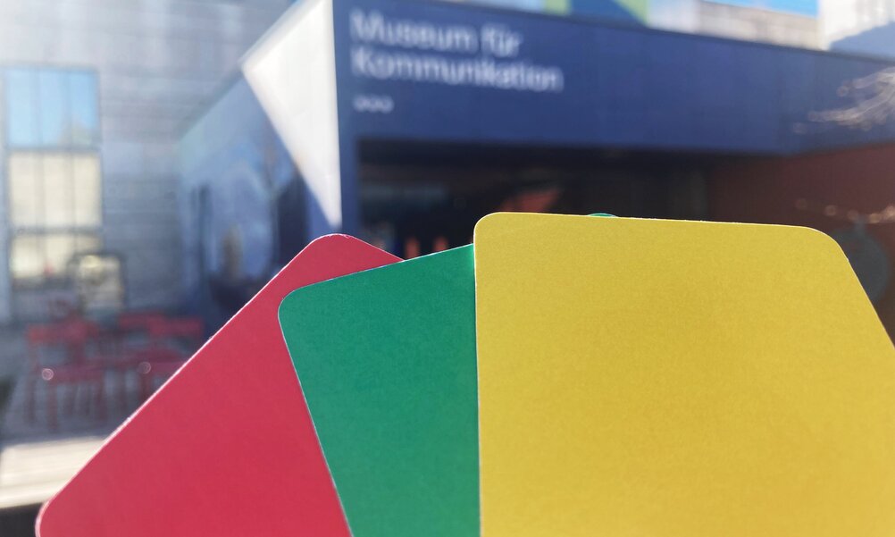 Drei farbige Karten in den Farben rot, grün und gelb vor dem Eingang des Museums für Kommunikation.