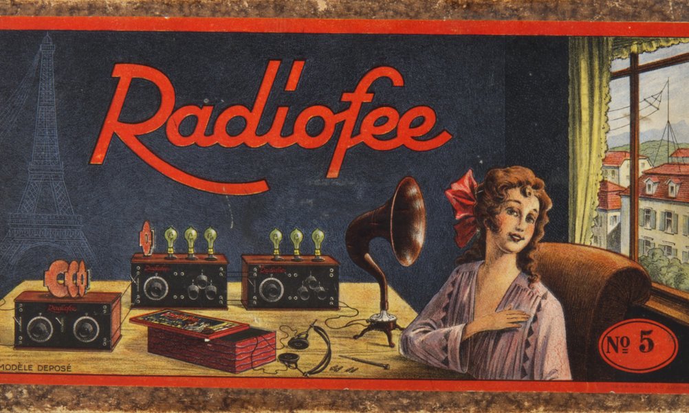 Abbildung einer Verpackungsschachtel für ein Radiogerät aus den 1920er-Jahren. Auf der Schachtel ist eine Frau mit Kopfhörern zu sehen, hinter ihr auf dem Tisch technisches Material zum Radioempfang.