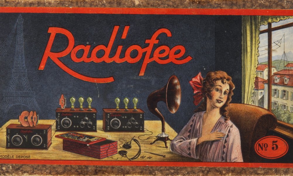 Abbildung einer Verpackungsschachtel für ein Radiogerät aus den 1920er-Jahren. Auf der Schachtel ist eine Frau mit Kopfhörern zu sehen, hinter ihr auf dem Tisch technisches Material zum Radioempfang.