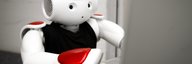 Man sieht einen weiss-roten Roboter in menschenähnlicher Gestalt. Man sieht sein Gesicht und seinen Oberkörper. Er sitzt vor einem Laptop. - vergrösserte Ansicht