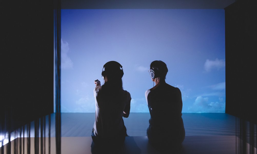 Two people sit in front of a projection of a sky with clouds.