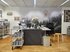 Blick in einen Arbeitsraum: Eine Arbeitsinsel mit Computern, Unterlagen und einem Blumenstrauss. Davor ein Handwagen mit Archivschachteln, hinten an der Wand ein raumhohes Bild eines Waldes im Nebel, daneben historische Bilder.