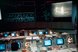 Blick ins nächtliche Kontrollzentrum der Apollo XI-Mission der NASA. Auf den Kontrollpulten leuchten Bildschirme, an der Wand ist eine Projektion zu sehen.