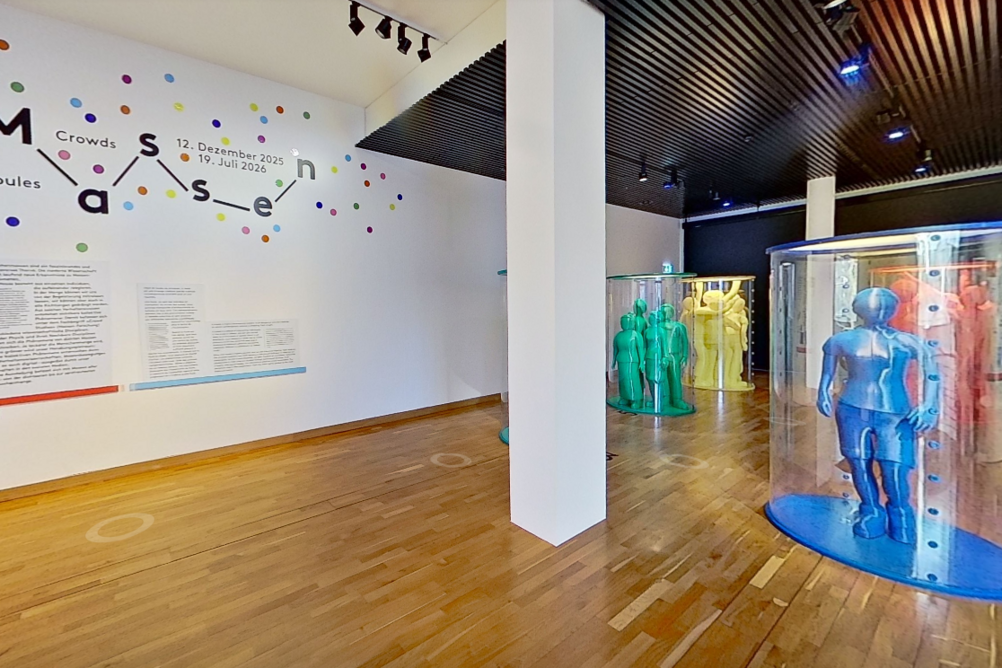Blick auf den Eingang der Ausstellung Massen. Links der Titel an der Wand, rechts farbige lebensgrosse Figuren in Plexiglassäulen.