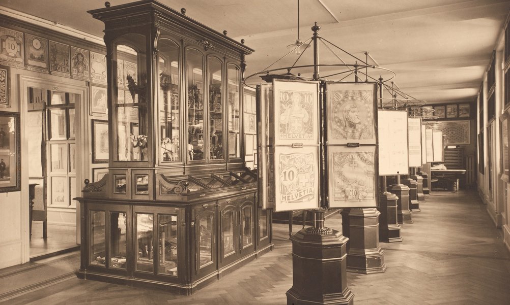 Sepia image of an old showroom. On display are enlarged stamps and, in an elegant display case, models of stagecoaches.