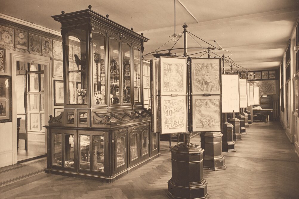 Sepia image of an old showroom. On display are enlarged stamps and, in an elegant display case, models of stagecoaches.