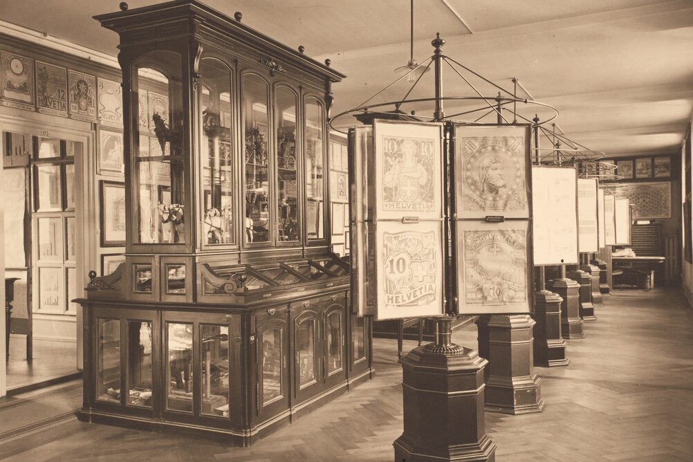 Museum historisch Sepiabild eines alten Ausstellungsraumes. Zu sehen sind vergrösserte Briefmarken und in einer eleganten Vitrine Modelle von Postkutschen.