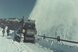 Auf der Farb-Fotografie aus der Zeit um 1940 bis 1950 ist eine schneebedeckte Strasse ersichtlich. Im Hintergrund befindet sich eine hügelige Landschaft und blauer Himmel. Im Vordergrund ist eine grosse, raupengetriebene Schneeschleuder im Einsatz. Vom Schleuderwerk wird eine massive Schneewolke über den rechten Strassenrand ausgeschleudert.