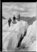 Schwarz-Weiss-Fotografie. Auf einem Gletscher drei wandernde Personen mit Hüten und mit Gehstöcken. Im Hintergrund Berge.