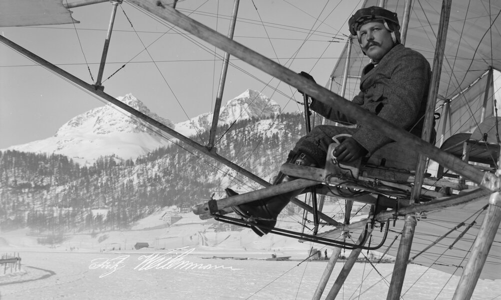 Schwarz-weiss Foto eines Flugpioniers. Er sitzt auf einem Konstrukt aus Stangen, Seilen und Tüchern in luftiger Höhe - hinter ihm sind die Alpen zu sehen.