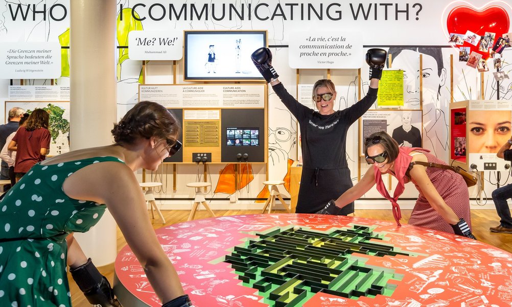 At a red gaming table, two women with dark glasses and boxing gloves steer a ball through a maze. A third woman cheers.