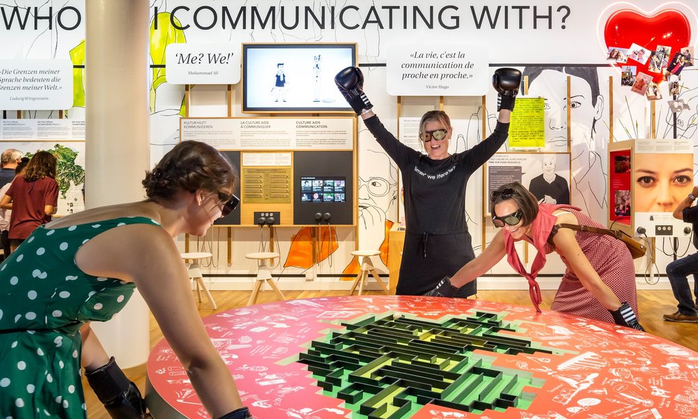 À une table de jeu rouge, deux femmes portant des lunettes noires et des gants de boxe contrôlent une balle dans un labyrinthe. Une troisième femme applaudit.