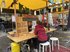 Sur un étal de marché avec un auvent jaune, deux personnes se parlent en s'asseyant.