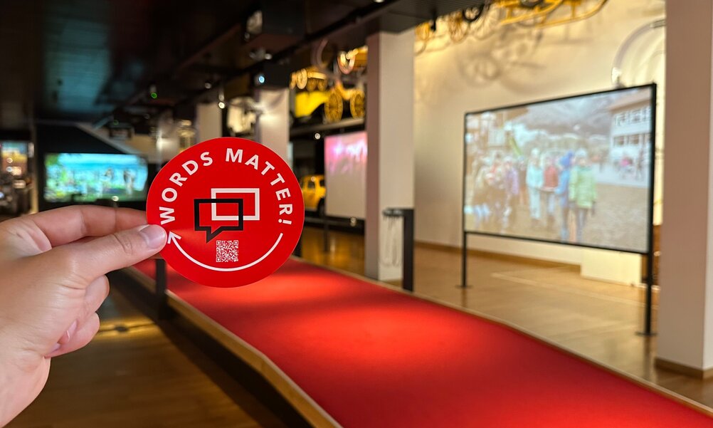 A hand holds a red sticker with the words ‘Words matter’ in the picture. An exhibition can be seen in the background.