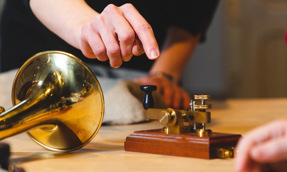 Senden Ein Posthorn und ein Morsegerät stehen auf einem Tisch, eine Hand ist gerade im Begriff, auf den Morsetaster zu drücken.