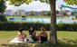 Sur une pelouse, sur une couverture sous un arbre, trois femmes sont assises et parlent entre elles. On aperçoit le bassin de la piscine extérieure à l'arrière-plan.