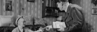 Historische Aufnahme eines Postboten, der in der Stube einer sitzenden Frau ein Couvert übergibt. Im Hintergrund Blumentapete, Bilderrahmen und Dekoration. - vergrösserte Ansicht