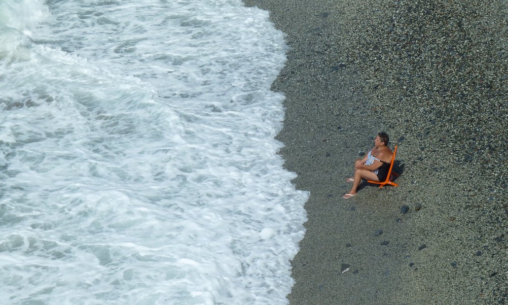 Eine Frau sitzt entspannt auf einem Liegestuhl am Strand, vor ihren Füssen tost die Brandung des aufgewühlten Meeres.