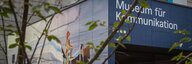 Durch die Blätter und Äste eines Baumes sieht man den Eingang des Museums für Kommunikation. Rechts die Beschriftung mit dem Museumsnamen, links ein Wandgemälde mit einer Biene in einer Flaschenpost, die übers Meer gleitet. - vergrösserte Ansicht