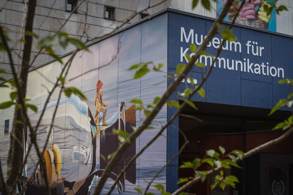 Durch die Blätter und Äste eines Baumes sieht man den Eingang des Museums für Kommunikation. Rechts die Beschriftung mit dem Museumsnamen, links ein Wandgemälde mit einer Biene in einer Flaschenpost, die übers Meer gleitet.
