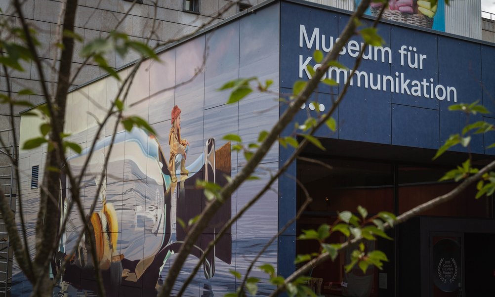 Durch die Blätter eines Gebüschs sieht man den Eingang des Museums für Kommunikation mit einem grossen Wandbild.
