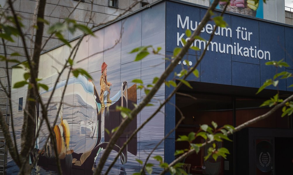 Through the leaves of a bush you can see the entrance of the Museum of Communication with a large mural.