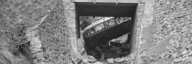 Historisches schwarz-weiss Bild. Blick durch ein Bachbett – diesmal bergaufwärts. Unter der Brücke durch sieht man das Postauto, das im Bachgraben steht. Darüber Menschen und dahinter Bäume. - vergrösserte Ansicht