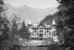 Historische schwarz-weiss Aufnahme: Zwischen Bäumen hindurch sieht man das Grandhotel Giessbach mit kleinen spitzen Türmchen. Im Hintergrund sind Berge zu sehen. - vergrösserte Ansicht