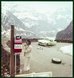 Historisches Farbfoto von einer Passstrasse in den Alpen. Eine Frau telefoniert im Vordergrund an einer SOS-Säule, im Hintergrund ein defektes Auto.