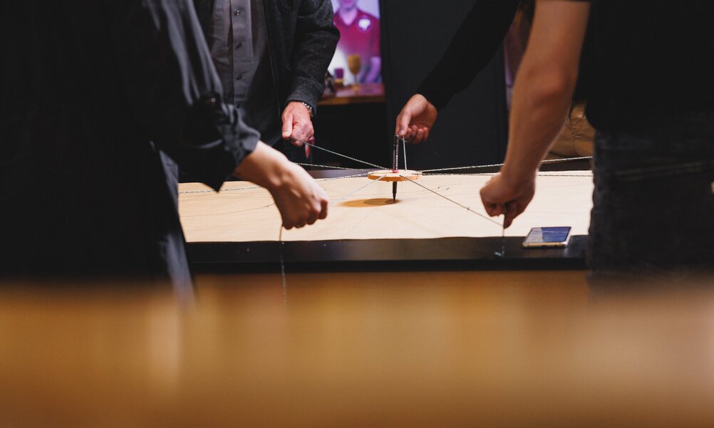 Vier Personen stehen um einen Tisch und halten Schnüre. Alle schnüre führen in die Mitte, dort ist ein Stift den alle Personen mit den Schnüren zusammen bewegen.