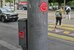 Ein grauer Pfosten einer Strassenlampe mit roten Klebern drauf, im Hintergrund Fussgänger:innen und Autos. - vergrösserte Ansicht