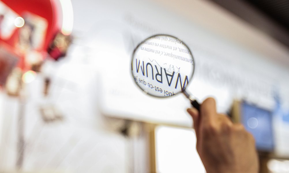 A hand holds a magnifying glass over the text "Why" in the exhibition.