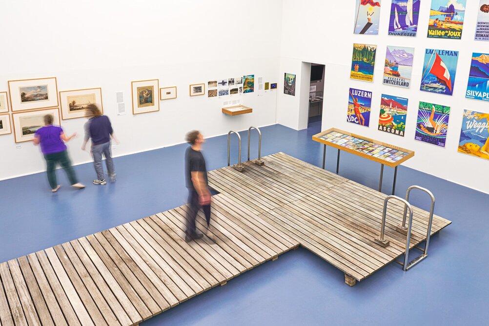 [Translate to English:] Personen in der Ausstellung auf einem inszenierten Steg, betrachten Poster und Grafiken von Schweizer Seen 