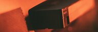 rötliches Bild mit Tiefenunschärfe: Eine Ecke eines Röhrenfernsehers mit Bildrauschen und darunter eine Videokassette, die halb im Gerät verschwindet. - vergrösserte Ansicht
