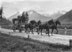 Historische Aufnahme einer Postkutsche mit vier Pferden im Galopp. Im Hintergrund der Landstrasse sind Berge zu sehen.