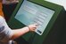 Eine Person steht vor einem Touchscreen und wählt darauf aus verschiedenen Handlungsbereichen ein Thema aus. - vergrösserte Ansicht