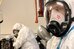 Foto von zwei Personen in Schutzkleidung - weisse Overalls und Atemmasken über dem ganzen Gesicht. Ein Person ist im Hintergrund an der Arbeit, die andere blickt durch die Maske in die Kamera. - vergrösserte Ansicht