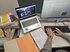 Schreibtisch mit Laptop, Bildschirm und Tastatur, darauf die Hände einer Person, die arbeitet. Auf dem Bildschirm ist ein Schwarzweiss-Foto zu sehen, neben der arbeitenden Person ein Ordner, Bücher, Archivschachteln.